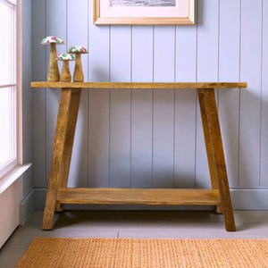 May include: A wooden console table with a natural finish, featuring a lower shelf and angled legs. Three decorative mushroom figurines with floral patterned caps sit on the table. The table is placed against a light blue wall.