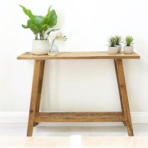 May include: A light wood console table with a lower shelf. The table has a simple, modern design with angled legs. There are three potted plants on the table, including a white ceramic pot with a green plant and two smaller pots with succulents.