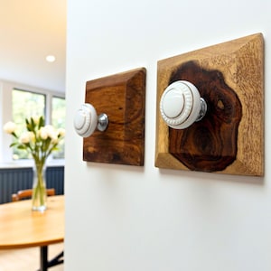 May include: Two wooden wall hooks with white ceramic knobs. The square wooden bases have a rich, dark brown color, and the knobs are adorned with a delicate silver trim. The hooks are mounted on a white wall, showcasing a blend of rustic and elegant design.