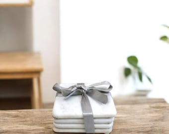 May include: Set of four white marble coasters with a gray ribbon tied around them. The coasters are on a wooden surface.