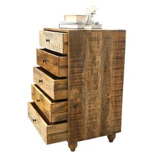 May include: A wooden chest of drawers with five drawers, some open. The chest is made of light brown wood with a distressed finish. The top drawer has a decorative carved design. A small vase of flowers and books sit on top.