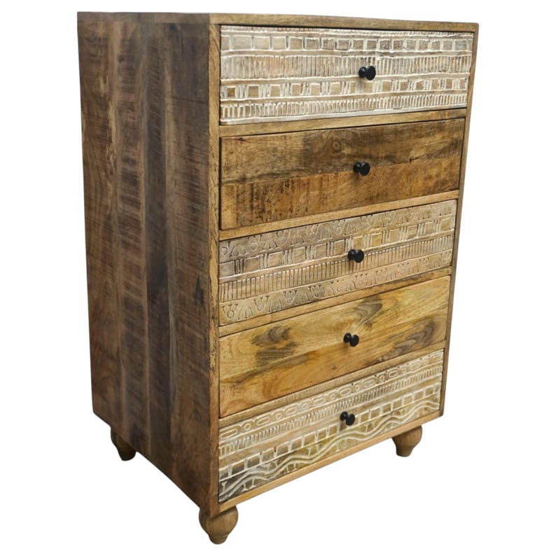 May include: A wooden chest of drawers with five drawers. The chest is made of light brown wood with a distressed finish. The top, middle, and bottom drawers have a carved white design. Each drawer has a small, round, black knob.