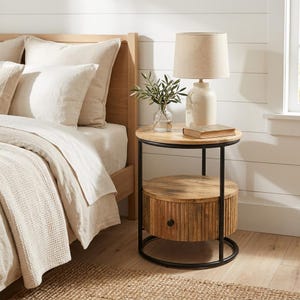 May include: A round wooden nightstand with a black metal frame. The nightstand has a top surface, a lower drawer, and a small lamp with a beige lampshade. A small vase with greenery and a book are on the top surface. The bed frame and bedding are visible in the background.