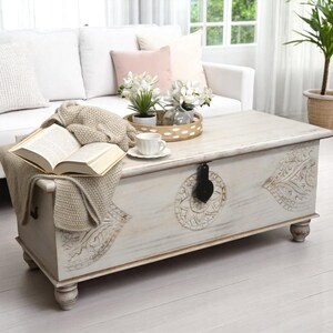May include: White wooden storage trunk with carved details, used as a coffee table in a living room setting. The trunk has a hinged lid and a black metal latch. A book, a knitted throw, and a cup and saucer are on top of the trunk.