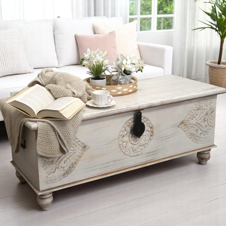 May include: White wooden storage trunk with carved details, used as a coffee table in a living room setting. The trunk has a hinged lid and a black metal latch. A book, a knitted throw, and a cup and saucer are on top of the trunk.