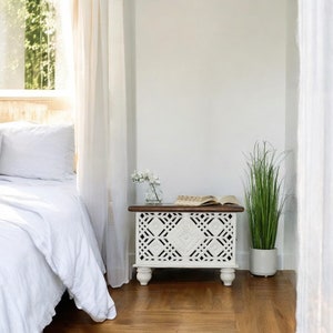May include: White wooden side table with intricate carved details and a brown top. The table has a book and a vase of white flowers on top. A potted plant is on the floor next to the table.