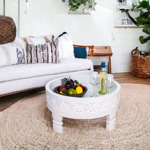 May include: A white round coffee table with carved details and four legs. The table is set with a bowl of fruit and two wine glasses. The table is on a natural fiber rug in a living room setting.