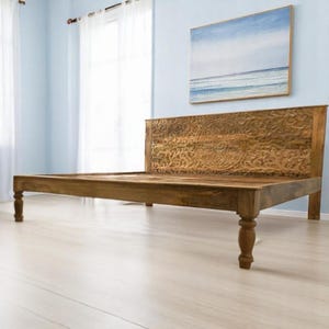 May include: A wooden bed frame with an intricately carved headboard. The bed frame is made of light brown wood and has four sturdy legs. The headboard features a detailed floral and geometric pattern. The bed frame is set against a light blue wall.
