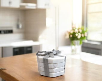 May include: Set of four white marble coasters with a gray ribbon tied around them. The coasters are hexagonal in shape and have a subtle marbled pattern.