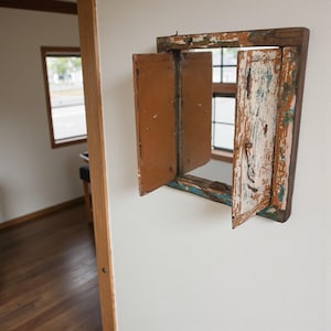 May include: A small, rustic wooden window frame with two hinged shutters, mounted on a white wall. The shutters are open, revealing a view of a window and the outside. The frame and shutters have a distressed, weathered appearance with shades of brown, blue, and white.