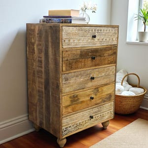 May include: A five-drawer wooden chest. The chest is made of light brown wood with a natural finish. The top and bottom drawers have a carved design. The drawers have black knobs and are set on short, rounded legs. The chest is approximately 100cm tall.