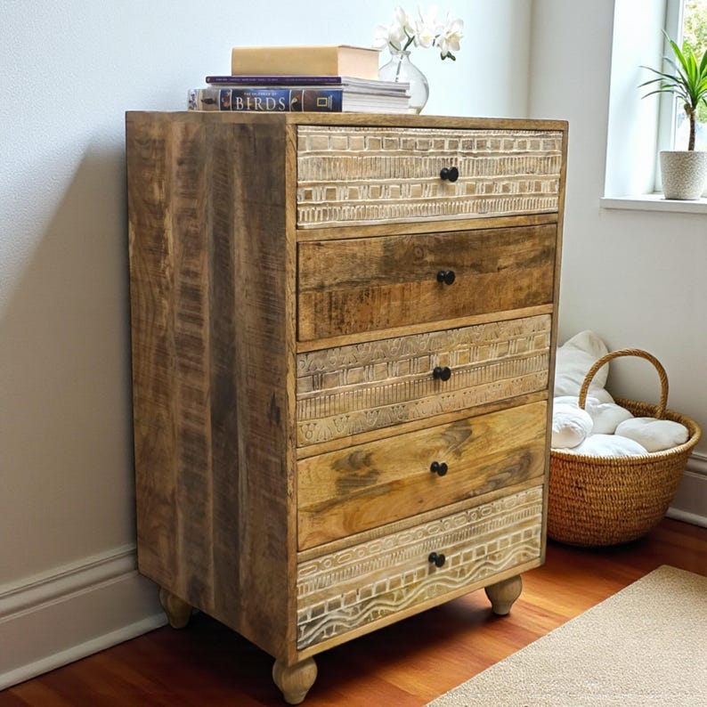 May include: A five-drawer wooden chest. The chest is made of light brown wood with a natural finish. The top and bottom drawers have a carved design. The drawers have black knobs and are set on short, rounded legs. The chest is approximately 100cm tall.