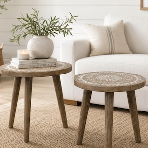 May include: Two small, round wooden side tables with a distressed finish. Each table has three angled legs and a decorative, carved design on the tabletop. One table holds a vase with greenery, books, and a candle. The tables are on a neutral rug.