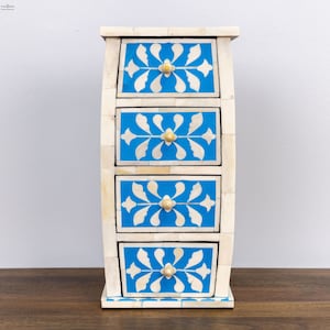 May include: A white bone inlay chest with four drawers. The drawers are painted a bright blue and have a white floral design. The chest has a curved front and sits on a wooden surface.