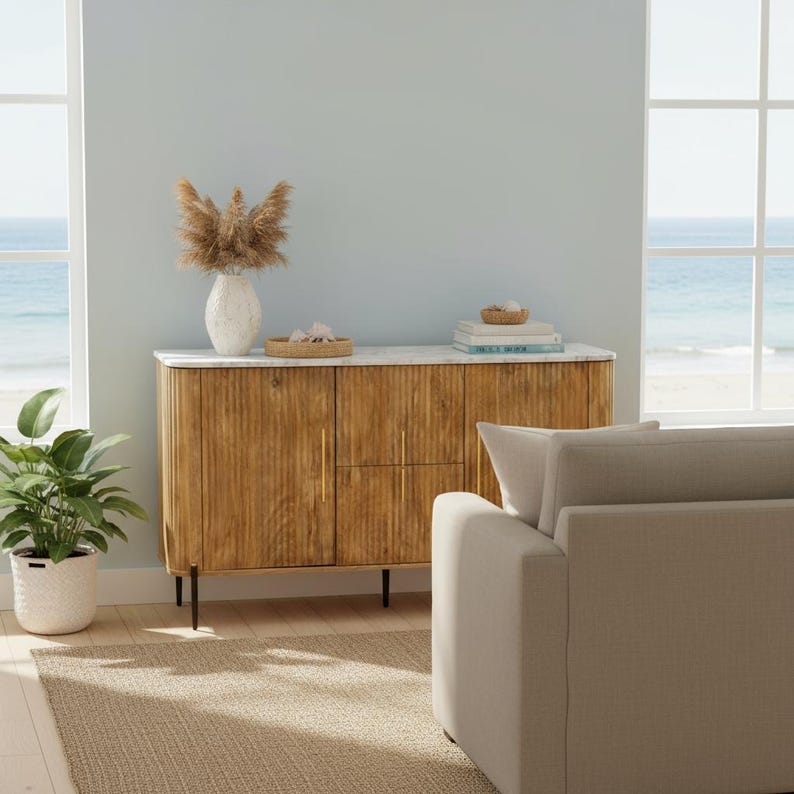 K&ouml;nnte beinhalten: Ein Holzschrank mit Marmorplatte und goldenen Griffen. Der Schrank befindet sich in einem Raum mit einem beigen Teppich, einer wei&szlig;en Vase mit getrockneten Blumen und einer gr&uuml;nen Pflanze in einem wei&szlig;en Topf. Der Hintergrund zeigt einen Strand und das Meer.