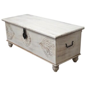 May include: White wooden storage chest with carved floral designs and a black metal handle. The chest has a hinged lid and sits on four turned legs.