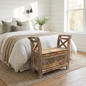 May include: A wooden storage bench with carved floral details sits at the foot of a bed. The bench has a distressed finish and a hinged lid. The bedroom features white bedding, neutral-toned pillows, and a woven rug, creating a cozy, rustic aesthetic.