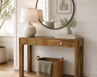 May include: A wooden console table with two drawers, a round mirror, and a lamp. A woven basket with a blanket sits below the table. A vase with greenery and books are on the table. The room has a neutral color palette.