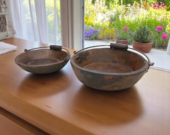 Könnte beinhalten: Zwei rustikale Metallschalen mit Griffen. Die Schalen sind rund mit dunkler, verwitterter Oberfläche und haben jeweils einen Holzgriff. Sie stehen auf einer Holzoberfläche, mit einer hellen Szene im Freien im Hintergrund.
