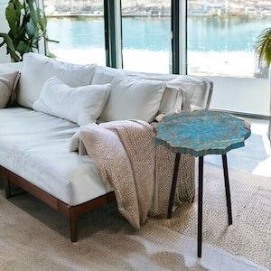 May include: A teal blue side table with a unique, intricate design on the top. The table has three black legs and is made of wood. The table is sitting on a light brown rug in front of a white couch.