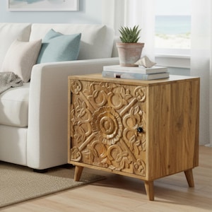 May include: A wooden side table with a carved floral design on the door. The light brown cabinet has a small black knob and sits on four angled legs. A small potted plant and books rest on top.