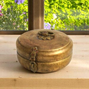 May include: A round, antique brass box with a hinged lid and ornate clasp. The box features a decorative handle and is set against a wooden surface near a window with a view of greenery.