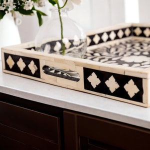 May include: A black and white bone inlay serving tray with a floral design and diamond-shaped accents. The tray has a handle and is sitting on a white countertop.