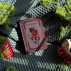 May include: A book titled "INSATIABLE" with a red and white cover featuring a heart and skull illustration. The book rests on a teal knitted blanket, surrounded by green moss and decorative elements, including a red candle and a skull.