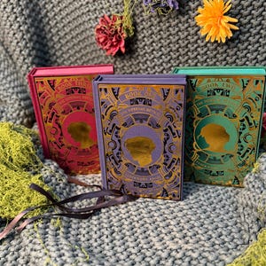 May include: Three hardcover books with ornate gold and purple designs. The books are in shades of pink, purple, and teal. The text on the book spines reads "Book One", "Book Two", and "Book Three". The books are on a gray knitted surface.