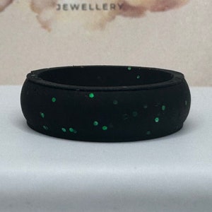 Op de afbeelding: Een zwarte ring met groene glitterstippen. De ring is een bandstijl, met een afgeronde vorm. De ring is gemaakt van een mat materiaal. De ring is een sieraad.