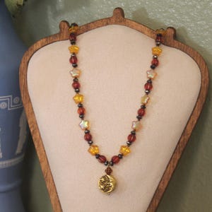 May include: A necklace featuring a circular gold-toned pendant with a celestial moon and star design. The chain consists of alternating dark red translucent beads, small metallic spacers, and iridescent star-shaped beads in shades of amber.