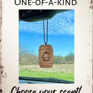 May include: A wooden barrel-shaped car air freshener with a black and white image of a barrel and the text "Bourbon Barrel" hanging from the rearview mirror of a car. The car is parked and the background is a blue sky with white clouds.