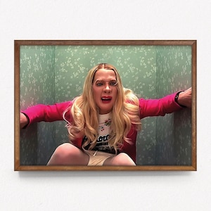 May include: A woman with blonde hair is wearing a pink jacket and a white shirt with a floral design. She is in a small room with green wallpaper and is looking at the camera with a surprised expression.