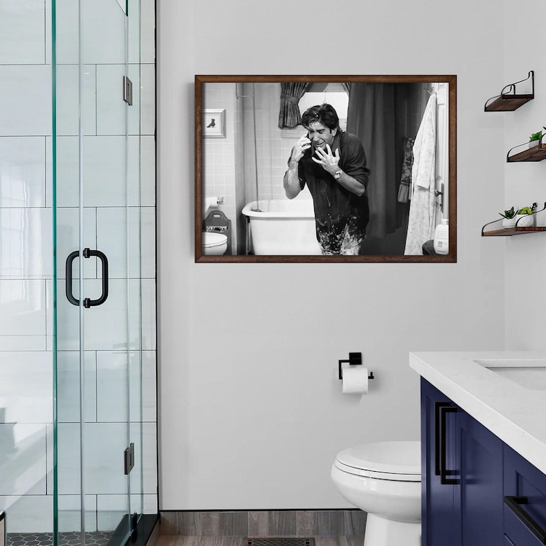 May include: A framed black and white photograph of a man in a bathroom, looking distressed while on the phone. The photograph is mounted on a white wall above a toilet and a blue vanity. A glass shower door is visible on the left.