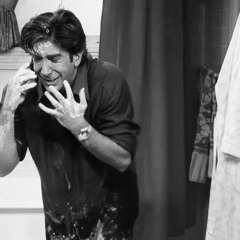 May include: Black and white image of a man in a dark shirt, covered in a white substance, talking on a phone. The man is smiling, with the white substance on his face, hands, and shirt. A watch is visible on his wrist.