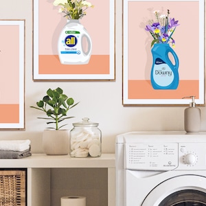 May include: Three framed prints with colorful floral arrangements in detergent bottles. The bottles are labeled "Tide", "All", and "Downy". The prints are hanging on a white wall above a white washing machine.
