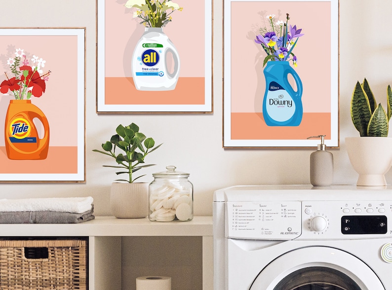 May include: Three framed prints with colorful floral arrangements in detergent bottles. The bottles are labeled "Tide", "All", and "Downy". The prints are hanging on a white wall above a white washing machine.