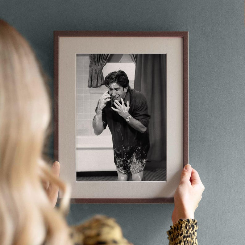 Peut inclure: Photographie encadr&eacute;e en noir et blanc d'un homme dans une salle de bain, semblant boulevers&eacute; au t&eacute;l&eacute;phone. Le cadre marron avec un passe-partout blanc est tenu par une personne aux cheveux blonds et une manche &agrave; motifs.