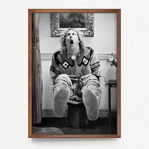 May include: Black and white framed print featuring a person with their mouth open, wearing a patterned shirt and fringed pants. The person is seated on a stool. A painting hangs on the wall behind them.