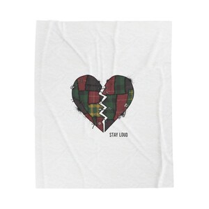 Könnte beinhalten: Eine weiße Decke mit einem verwitterten Herzdesign. Das Herz ist gespalten, mit kariertem Stoff in Rot, Grün und Gelb geflickt und mit Sicherheitsnadeln zusammengehalten. Die Worte "STAY LOUD" sind darunter gedruckt.