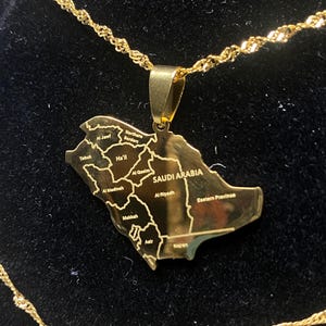 May include: A gold-coloured pendant necklace depicting a detailed map of Saudi Arabia. The pendant includes the country's name and regional names. The chain has a twisted rope design.