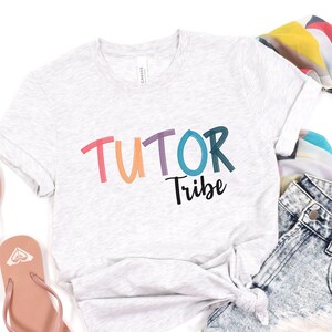 May include: A light gray t-shirt with the text "TUTOR Tribe" in rainbow colors. The text is in a playful, hand-drawn style.