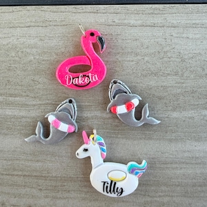 Puede incluir: Cuatro dijes decorativos que incluyen un flotador de flamenco rosa brillante con el nombre Dakota, dos tiburones grises con salvavidas rojos y blancos, y un flotador de unicornio blanco con crin y cola coloridas con el nombre Tilly en letra negra.