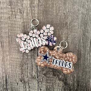 Op de afbeelding: Twee doorzichtige sleutelhangers in de vorm van een hondenbot, met een Dallas-thema. De ene sleutelhanger heeft een wit baseballpatroon met een blauw New York Yankees-logo en het woord "Dallas". De andere sleutelhanger heeft een bruin voetbalpatroon met een blauwe Dallas Cowboys-ster en het woord "Dallas".