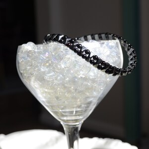 May include: A black beaded bracelet with silver accents sits on top of a clear glass bowl filled with iridescent glass beads.