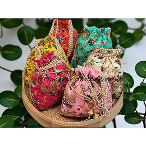 以下が含まれることがあります： 赤、黄、ピンク、ティール、クリーム色の刺繍入り巾着袋のコレクション。各ポーチは花柄で、ビーズのドローストリングが付いています。ポーチは木製のトレイに配置され、「Grow handmade in india」の文字が表示されています。