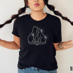 Può includere: T-shirt nero con un design di strass di due gatti. I gatti sono delineati con piccoli strass scintillanti. La maglietta ha un girocollo classico e maniche corte. Il testo "SS10" si trova nell'angolo in basso a destra.