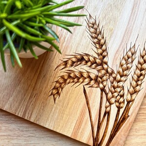 May include: A wooden chopping board with a design of wheat stalks burned into the surface. The board is light brown with a natural wood grain.