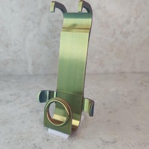 May include: A metallic green and gold iridescent stand featuring a vertical back panel with two top hooks and a circular base support with two side arms. The structure is constructed from a single piece of bent material and rests on a small white base.