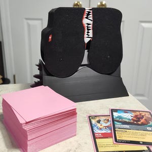 Puede incluir: Un porta tarjetas negro con un diseño de boca de monstruo rojo y blanco. El porta tarjetas está sentado en una mesa con una pila de tarjetas rosas y dos cartas coleccionables. Las cartas coleccionables presentan ilustraciones de personajes del juego "Magic: The Gathering". El texto en las cartas dice "Pete, Cazador de Tesoros" y "Kakamora, Estrella de Mar y Pizza".